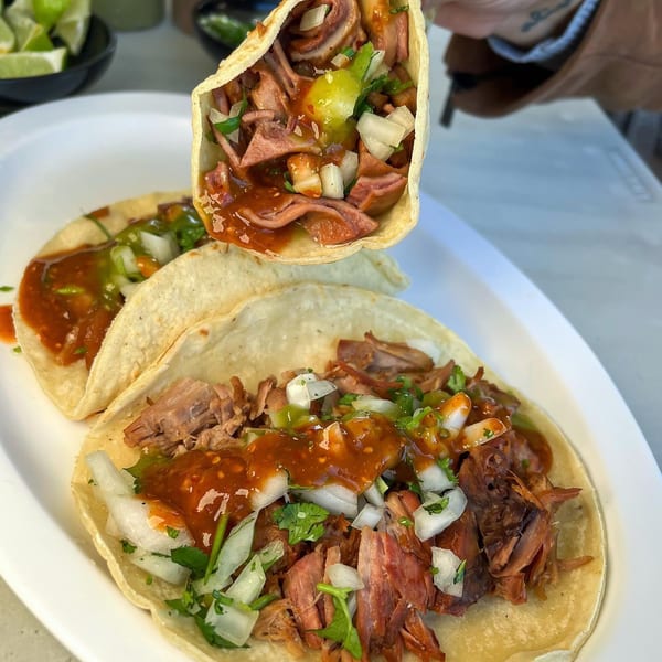 A hand holds up a taco filled with meat, onions, and salsa, with two more tacos on a plate.