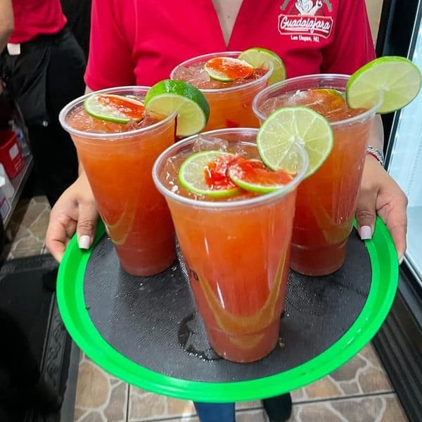 A server in a red shirt holds a tray with four red drinks garnished with lime slices.