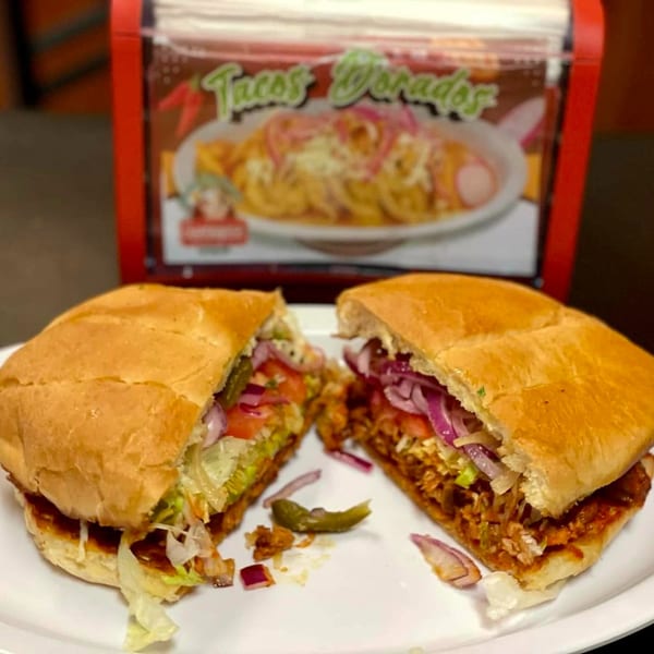 A large Mexican torta sandwich, cut in half, filled with meat, lettuce, tomato, and red onion.