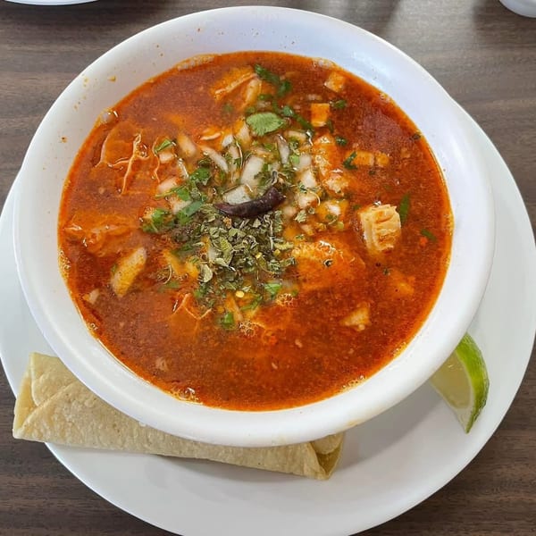 A bowl of menudo soup with cilantro, onions, and a dried chili, served with a tortilla and lime.