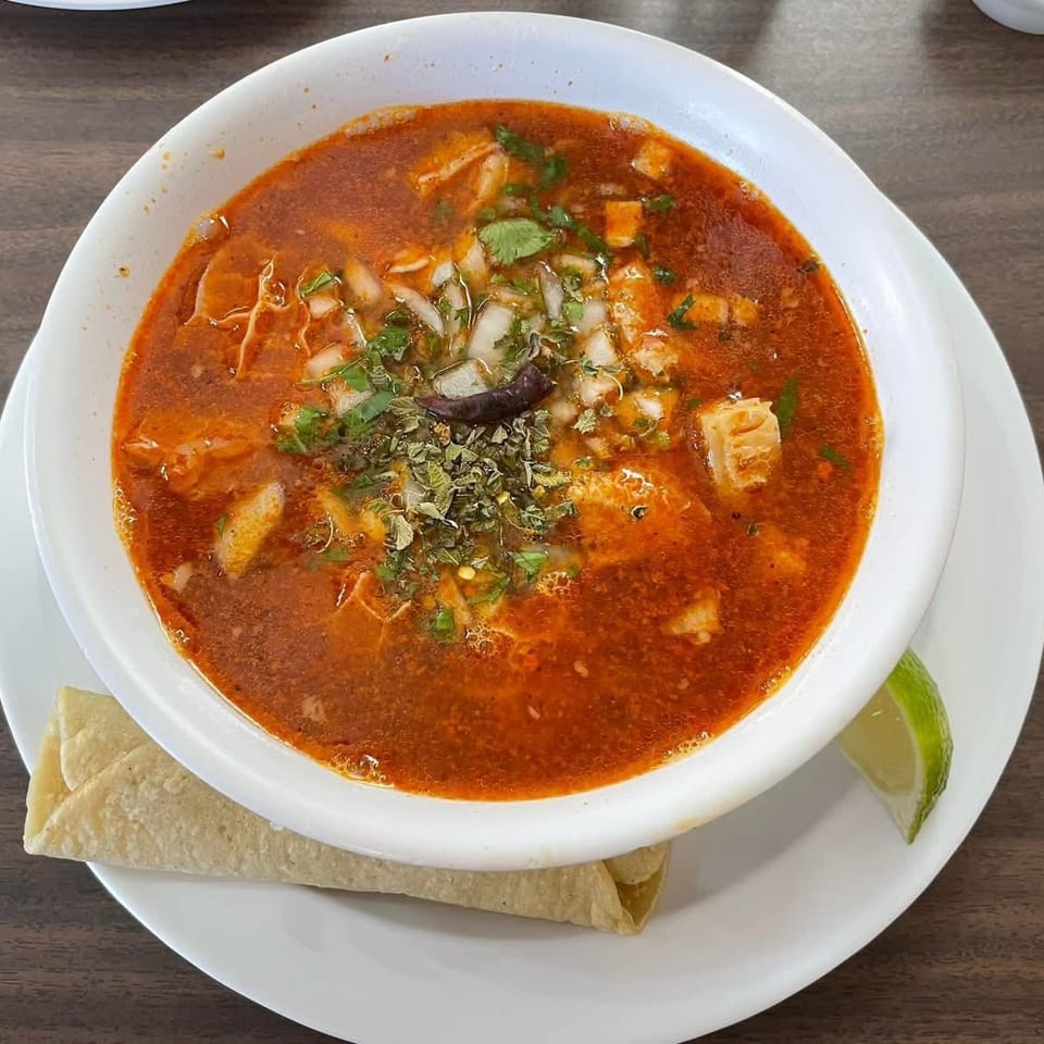 A bowl of menudo soup with cilantro, onions, and a dried chili, served with a tortilla and lime.
