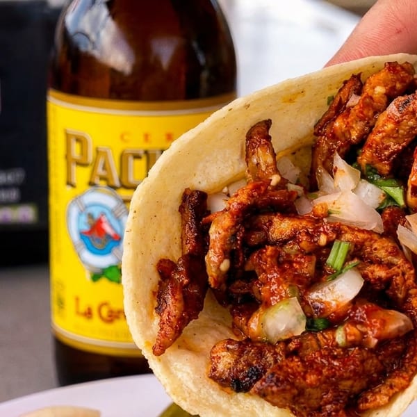 Hand holding a taco filled with marinated meat, onions, and cilantro, with a beer bottle in the background.