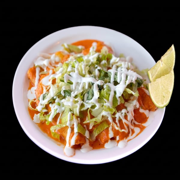 A plate of enchiladas topped with red sauce, shredded lettuce, sour cream, and lime wedges.